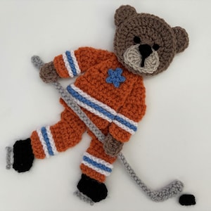 May include: A crocheted brown bear wearing an orange and blue hockey jersey and holding a grey hockey stick. The bear is on a white background.