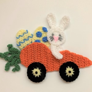 May include: A crocheted Easter bunny driving a carrot-shaped car. The bunny is white with gray whiskers and a pink nose. The car has black wheels and a yellow and blue egg in the back.