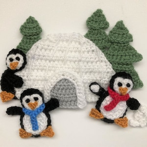 May include: Crocheted penguin ornaments with a white igloo and green trees. The penguins are wearing scarves and one is holding a snowball.