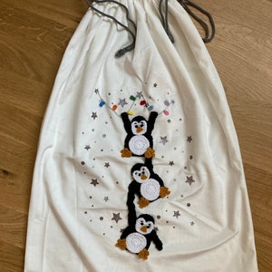 K&ouml;nnte beinhalten: Wei&szlig;e Stoffgeschenkt&uuml;te mit Kordelzug. Die Tasche zeigt ein Design mit drei geh&auml;kelten Pinguinen, die an einer Lichterkette mit Sternen im Hintergrund h&auml;ngen.