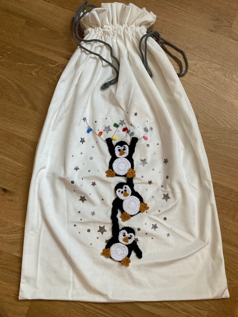 K&ouml;nnte beinhalten: Wei&szlig;e Stoffgeschenkt&uuml;te mit Kordelzug. Die Tasche zeigt ein Design mit drei geh&auml;kelten Pinguinen, die an einer Lichterkette mit Sternen im Hintergrund h&auml;ngen.