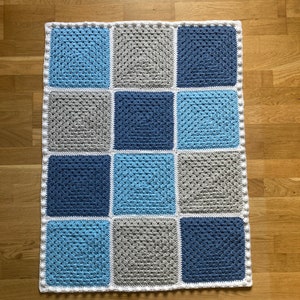 Op de afbeelding: Een gehaakt babydekentje met een witte rand en vierkanten in tinten blauw en grijs.