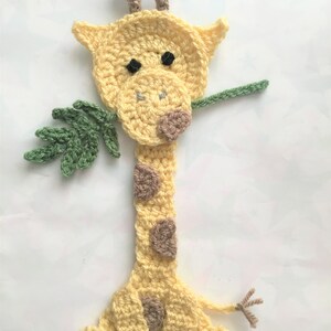 May include: A crocheted giraffe bookmark with brown spots and a green leaf in its mouth. The giraffe is yellow and has brown ears and a brown nose.