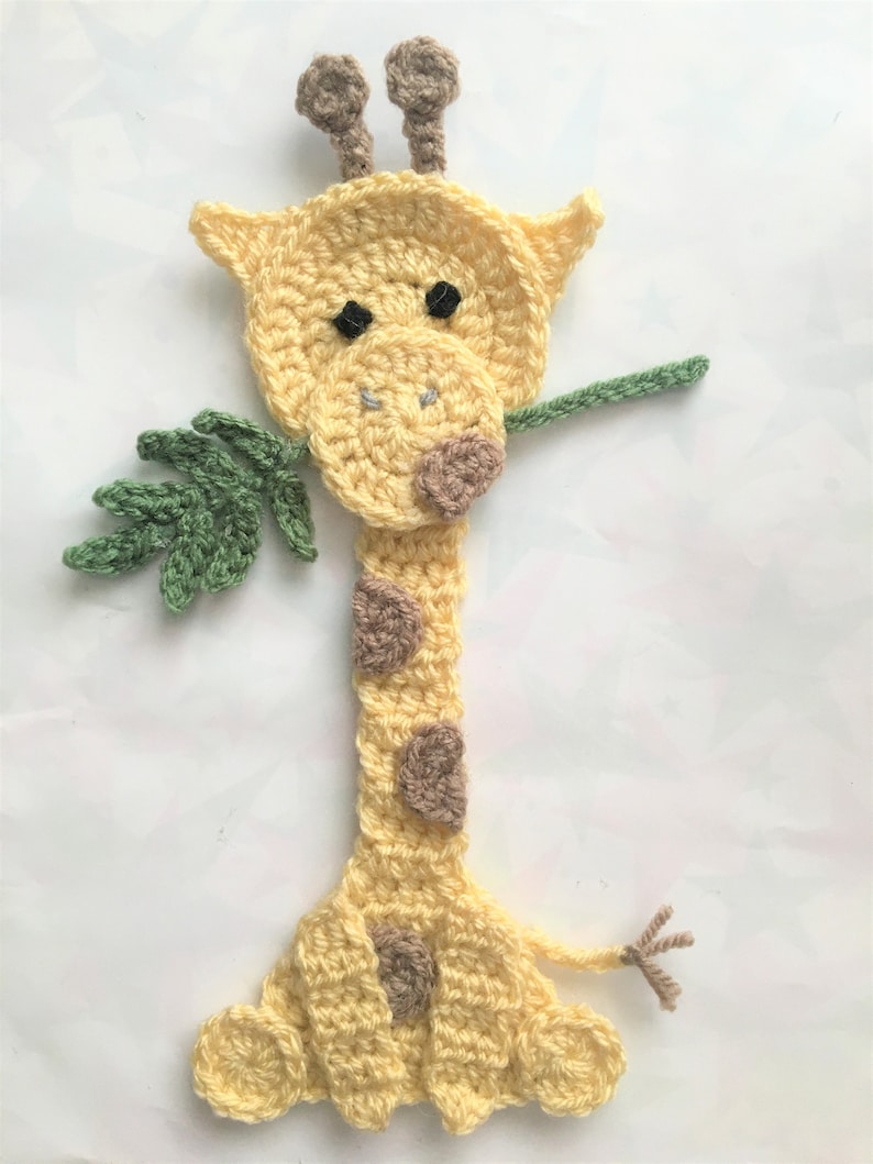 May include: A crocheted giraffe bookmark with brown spots and a green leaf in its mouth. The giraffe is yellow and has brown ears and a brown nose.