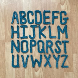May include: A set of blue crocheted letters spelling out the alphabet, A through Z, on a woodgrain background.