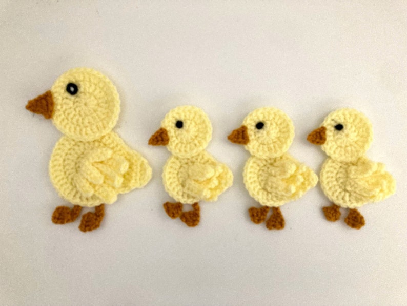 May include: Four crocheted yellow ducklings with brown beaks and black eyes. The ducklings are different sizes and are arranged in a row.