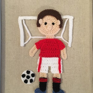 May include: Crocheted soccer player with a red jersey, white shorts, and black socks. The player is lying on a white crocheted goal with a black and white soccer ball in front of them.