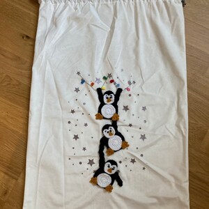 K&ouml;nnte beinhalten: Wei&szlig;e Stoffgeschenkt&uuml;te mit Kordelzug. Die Tasche zeigt ein gesticktes Design mit drei Pinguinen, die an einer Lichterkette h&auml;ngen, vor einem Hintergrund aus silbernen Sternen.