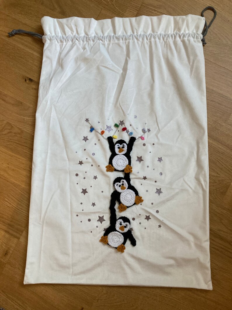 K&ouml;nnte beinhalten: Wei&szlig;e Stoffgeschenkt&uuml;te mit Kordelzug. Die Tasche zeigt ein gesticktes Design mit drei Pinguinen, die an einer Lichterkette h&auml;ngen, vor einem Hintergrund aus silbernen Sternen.