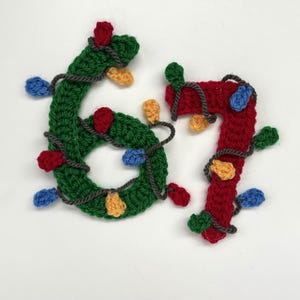 Peut inclure: Ornement de Noël crocheté représentant les chiffres six et sept. Le chiffre six est vert et le chiffre sept est rouge. Les deux sont ornés de lumières colorées rouges, bleues et jaunes, et enveloppés de fil gris.