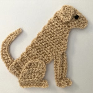 Könnte beinhalten: Gehäkelte Hundeapplikation in sitzender Position. Der Hund ist aus beigefarbenem Garn mit schwarzen Augen gefertigt. Der Schwanz ist eingerollt und die Ohren sind schlaff. Dieser handgefertigte Artikel eignet sich für Bastelarbeiten und Verzierungen.