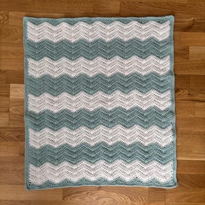 Może przedstawiać: Ręcznie robiony koc szydełkowy ze wzorem w zygzaki. Koc ma naprzemienne rzędy białej i turkusowej przędzy. Prostokątny koc wydaje się być wykonany z miękkiej przędzy. Koc jest wystawiony na drewnianej podłodze.