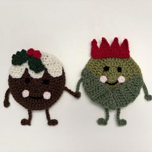 May include: Two crocheted Christmas ornaments. One is a brown pudding with white icing, green holly, and red berries. The other is a green and brown ornament with a red crown. Both have smiling faces and hold hands.