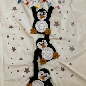 K&ouml;nnte beinhalten: Drei geh&auml;kelte Pinguine h&auml;ngen an einer Lichterkette mit bunten Lichtern vor einem wei&szlig;en Hintergrund mit silbernen Glitzersternen.