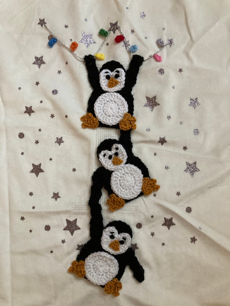 K&ouml;nnte beinhalten: Drei geh&auml;kelte Pinguine h&auml;ngen an einer Lichterkette mit bunten Lichtern vor einem wei&szlig;en Hintergrund mit silbernen Glitzersternen.