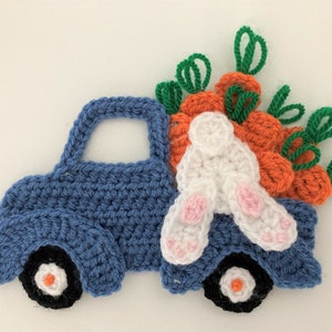 May include: A blue crocheted toy truck carrying a white crocheted bunny and orange crocheted carrots with green tops.