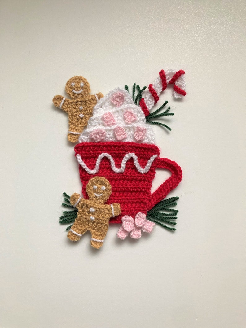 May include: Crocheted Christmas ornament featuring a red mug with white whipped cream and a candy cane, surrounded by gingerbread men and pink flowers.