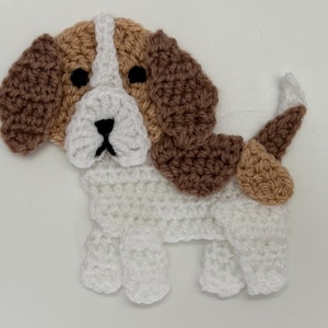 Puede incluir: Aplicación de perro de ganchillo en blanco, marrón y beige. El perro tiene orejas marrones, una cara beige y blanca y un cuerpo blanco. La cola del perro es beige y blanca. La aplicación está hecha de hilo.