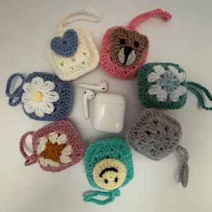 May include: A collection of handmade crochet AirPod cases in various colors and designs. The cases feature floral, heart, and bear motifs. A white AirPod case and earbuds are in the center. Each case has a loop for easy carrying.