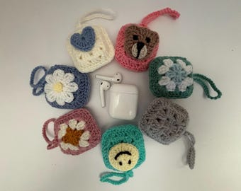 May include: A collection of handmade crochet AirPod cases in various colors and designs. The cases feature floral, heart, and bear motifs. A white AirPod case and earbuds are in the center. Each case has a loop for easy carrying.