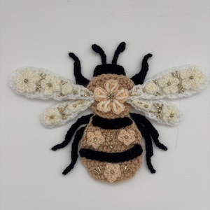 Op de afbeelding: Een handgemaakte gehaakte bijenapplicatie. De bij heeft een bruin lichaam met zwarte strepen, crèmekleurige vleugels versierd met kleine bloemen en zwarte antennes. Gouden accenten geven het ontwerp een vleugje glans.