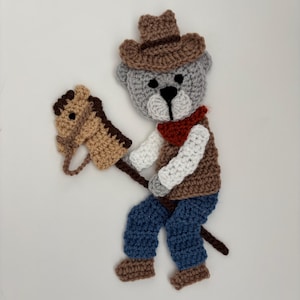 May include: A crocheted applique of a gray bear wearing a brown cowboy hat, a red bandana, a brown vest over white sleeves, and blue pants. The bear is holding a tan hobby horse with a dark brown mane and bridle on a dark brown stick.