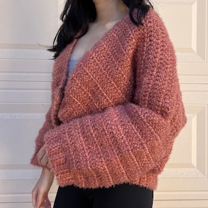 May include: A soft, pink cardigan with a V-neck and long sleeves. The cardigan is made of a fuzzy yarn and has a relaxed fit.