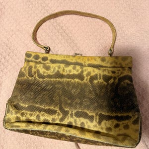 Puede incluir: Un bolso de mano estructurado con un patrón texturizado similar a la piel de serpiente en tonos marrón y beige. El bolso tiene un asa superior, un cuerpo rectangular que se estrecha ligeramente hacia arriba y un pequeño cierre metálico central.