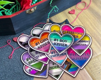 Op de afbeelding: Een verzameling hartvormige stickers met verschillende regenboogvlagontwerpen en tekst, waaronder "#1 Girl Kisser", "#1 Boy Kisser", "#1 Everyone Kisser" en "#1 People Kisser".