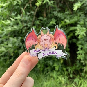 May include: A clear acrylic charm with a pink and red cartoon monster with a third eye and wings. The monster is holding a banner that says "Monster Fucker".