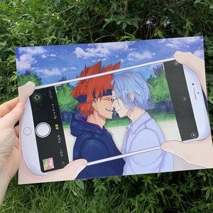 Op de afbeelding: Een telefoonscherm toont een digitale illustratie van twee anime-personages, een met rood haar en de ander met blauw haar, die elkaar aanlachen. De telefoon wordt vastgehouden door een hand.