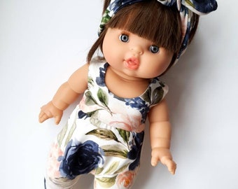 May include: A doll wearing a white and blue floral jumpsuit with a matching headband. The doll has brown hair, blue eyes, and a pink mouth.