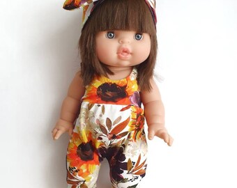 May include: A doll wearing a floral print jumpsuit and a matching headband. The doll has brown hair, blue eyes, and a pink mouth.