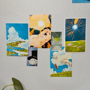 May include: A collection of colorful art prints featuring various scenes. One print depicts a blue sky with clouds and a landscape. Another shows a hand holding a bright light. Other prints include a cartoon character and a field with a bright light.