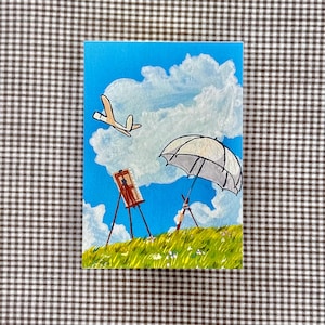 May include: Acrylic painting on canvas depicting a sunny landscape with a blue sky, fluffy white clouds, and green grass. The scene includes a white umbrella, an easel with a painting, and a cartoon airplane. The painting is on a rectangular canvas.