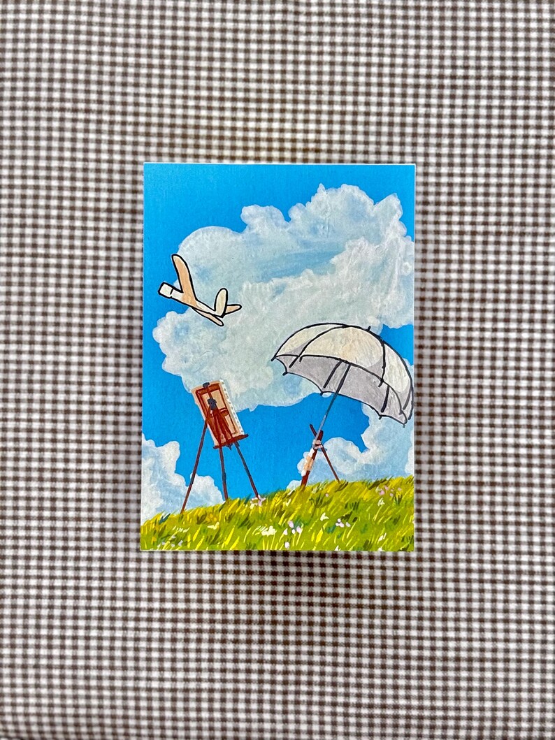 May include: Acrylic painting on canvas depicting a sunny landscape with a blue sky, fluffy white clouds, and green grass. The scene includes a white umbrella, an easel with a painting, and a cartoon airplane. The painting is on a rectangular canvas.