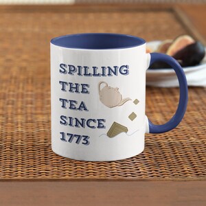 Op de afbeelding: Witte keramische mok met een blauwe binnenkant en handvat. De mok heeft de tekst "Spilling the Tea Since 1773" met een theepot illustratie. De mok staat op een geweven oppervlak.