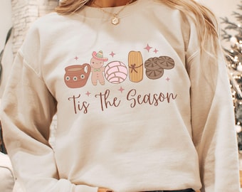 May include: A light beige sweatshirt with a festive design featuring a variety of Mexican pastries and the text "Tis the Season".