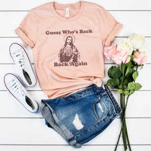 May include: A peach-colored t-shirt with a vintage-style image of Jesus and the text "Guess Who's Back Back Again".