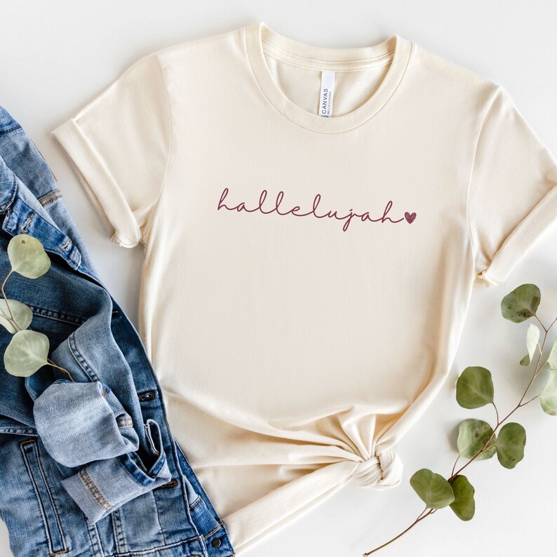 May include: A cream colored t-shirt with the word "hallelujah" written in maroon script with a small maroon heart to the right of the word.