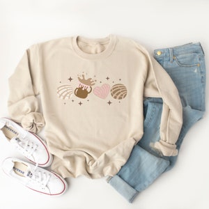 May include: A beige sweatshirt with a graphic design of a brown mug with a splash of chocolate, a pink heart-shaped pastry, and a brown pastry with a swirl design. The graphic is surrounded by small gold stars.