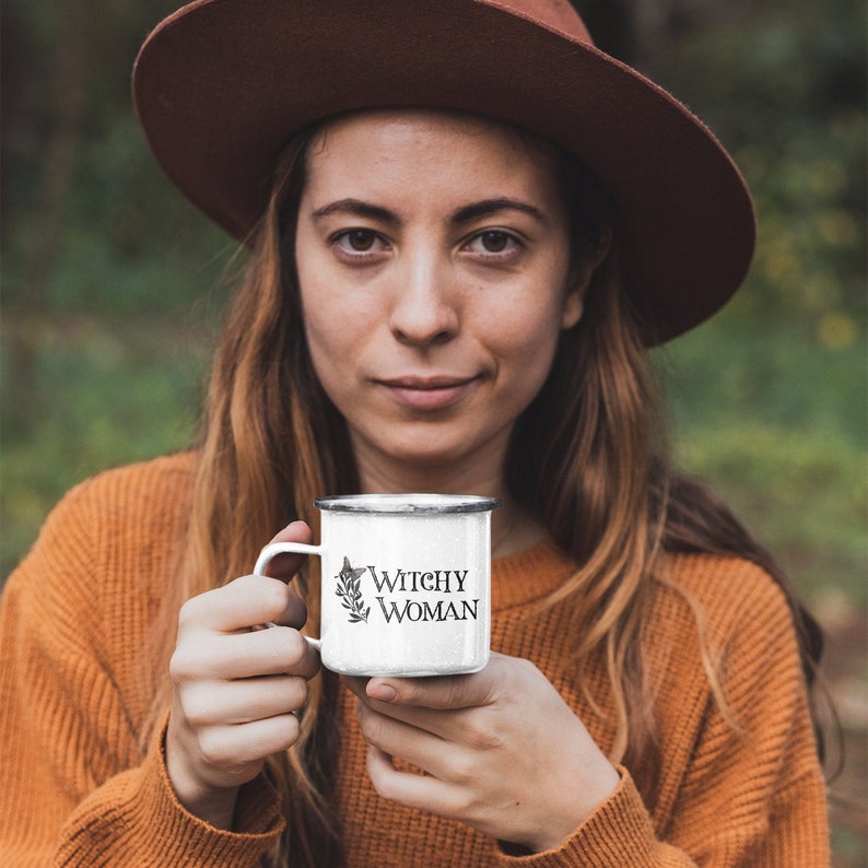 Pode incluir: Uma caneca de esmalte branco com um gr&aacute;fico preto e branco de uma bruxa e o texto "Witchy Woman". A caneca est&aacute; sendo segurada por uma pessoa que est&aacute; usando um chap&eacute;u marrom e um su&eacute;ter laranja.