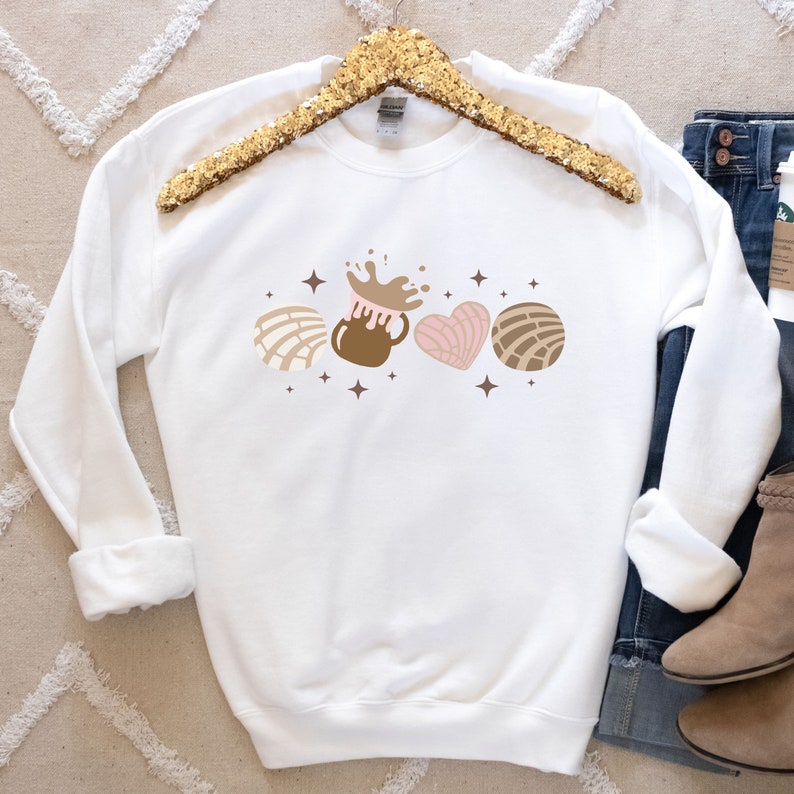 May include: White sweatshirt with a brown, pink, and white graphic of a concha, a heart-shaped concha, and a cup of hot chocolate with a splash of chocolate on top. The graphic is surrounded by small brown stars.