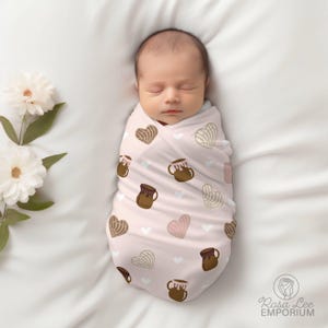 May include: A pink swaddle blanket with a pattern of brown hearts, brown mugs, and white hearts. A baby is sleeping inside the swaddle.