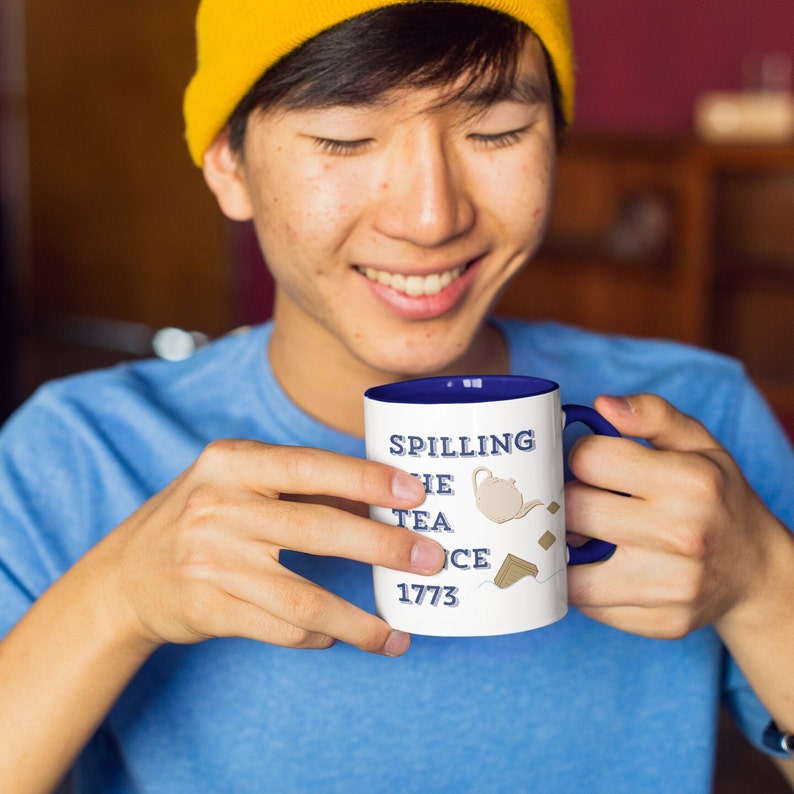 Op de afbeelding: Witte keramische mok met een blauwe binnenkant en handvat, met de tekst "Spilling the Tea Since 1773" met theepot illustraties. De mok wordt vastgehouden door een persoon die een blauw shirt en een gele muts draagt.