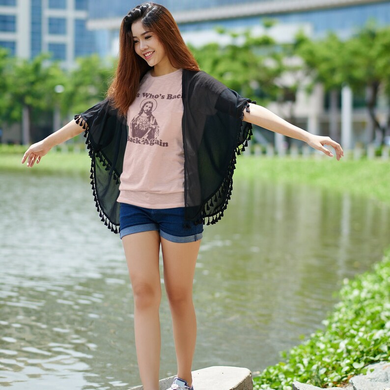 May include: A woman wearing a light pink t-shirt with the text "He's Who's Back Again" and a black sheer cardigan with tassels. She is standing in front of a body of water.