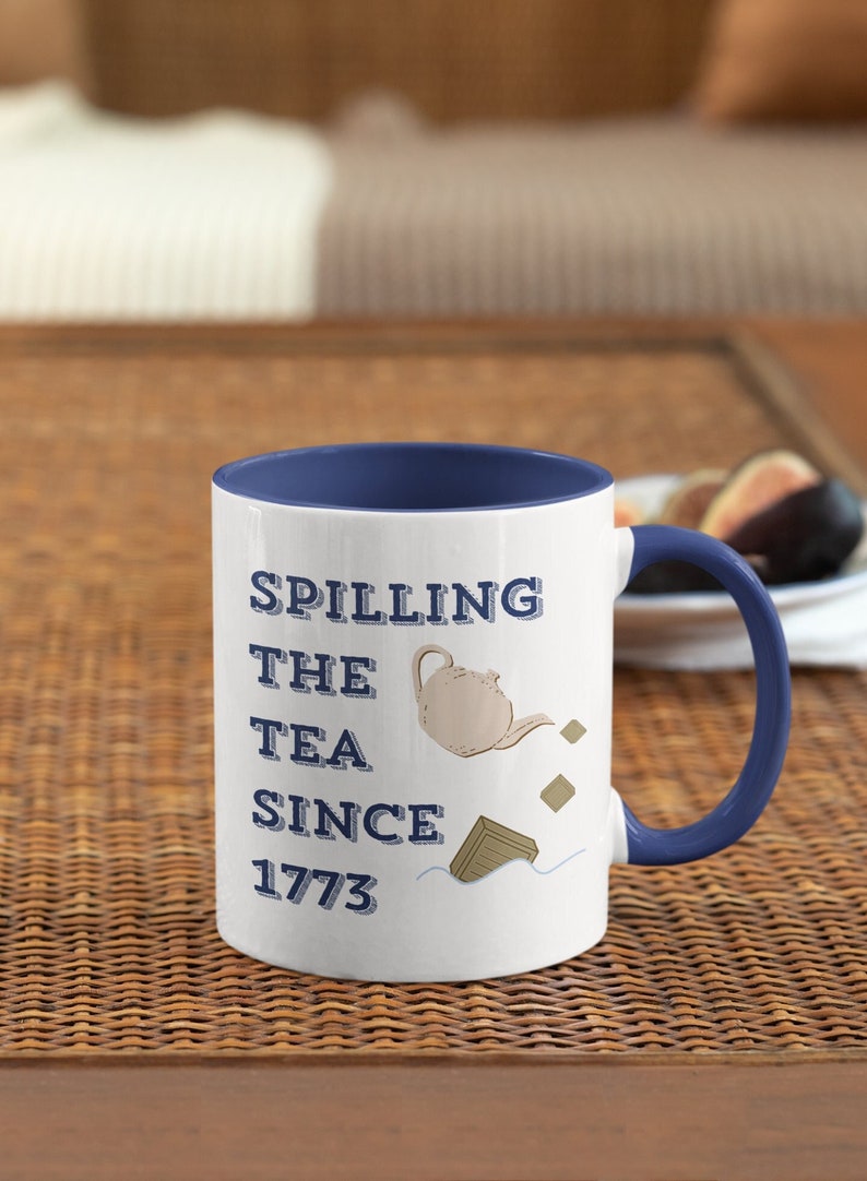 Op de afbeelding: Witte keramische mok met een blauwe binnenkant en handvat. De mok heeft de tekst "Spilling the Tea Since 1773" met een theepot illustratie. De mok staat op een geweven oppervlak.