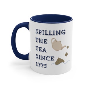 Op de afbeelding: Witte keramische mok met een marineblauwe handgreep en binnenkant. De mok heeft de tekst "Spilling the Tea Since 1773" met een illustratie van een theepot en theebladeren. Een leuk cadeau.