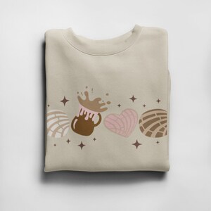 May include: A beige sweatshirt with a brown, pink, and white design of a mug of hot chocolate with a splash of chocolate, a heart-shaped pastry, and two concha pastries. The design is surrounded by small stars.