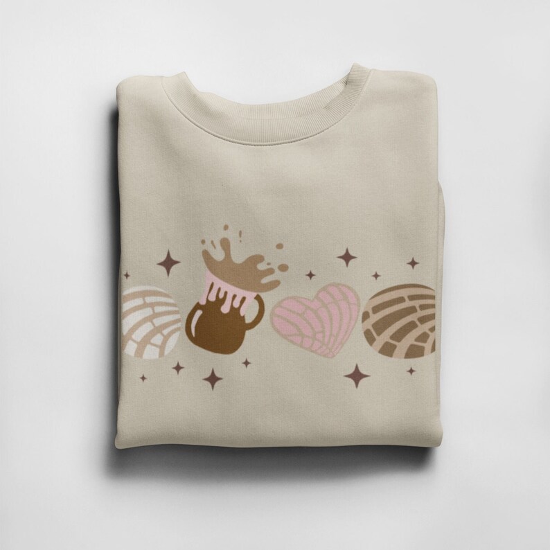 May include: A beige sweatshirt with a brown, pink, and white design of a mug of hot chocolate with a splash of chocolate, a heart-shaped pastry, and two concha pastries. The design is surrounded by small stars.